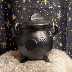 May include: A black cast iron cauldron with a matching lid, featuring a triple moon symbol embossed on the front. The pot has three short legs, two side handles with silver-toned metal rings, and a rounded body with a textured finish.