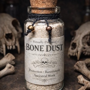 May include: A glass jar filled with bone dust, sealed with a cork stopper. The jar is labeled "Ritually Prepared BONE DUST" with a skull and crossbones graphic. The label also includes the words "Protection, Banishment, Ancestral Work."