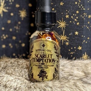 May include: A clear glass dropper bottle containing liquid and dried botanicals. The label is gold with black text reading RITUAL OIL SCARLET TEMPTATION ATTRACTION TEMPTATION SEDUCTION, featuring a rose graphic and decorative borders.