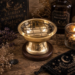 Puede incluir: Un quemador de incienso de latón pulido con una rejilla metálica extraíble y un símbolo de triple luna grabado en la base. El recipiente en forma de cuenco descansa sobre una base circular de madera oscura, diseñado para sostener incienso.
