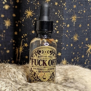 May include: A clear glass dropper bottle with a black cap and a gold label. The label features black text reading RITUAL OIL FUCK OFF BOUNDARIES REMOVAL BANISHING, along with a central illustration of a hand making a middle finger gesture.