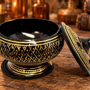 May include: A black bowl with a matching lid, featuring a gold-colored lattice pattern etched into the exterior. The bowl has a pedestal base and a handle on the lid. The interior of the bowl is smooth and black, while the exterior is decorated with gold lines.
