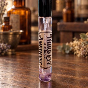 May include: A clear glass spray bottle with a black cap containing liquid and small purple stones at the base. A tan label on the front features the text Third Eye Amethyst with a moon phase graphic. The bottle is cylindrical and transparent.