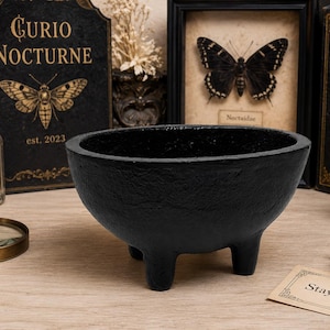 May include: A matte black, round, three-legged bowl with a textured surface. The bowl has a wide, open top and a deep basin, standing on three short, sturdy feet. The exterior finish is rough and dark, providing a solid, heavy appearance.