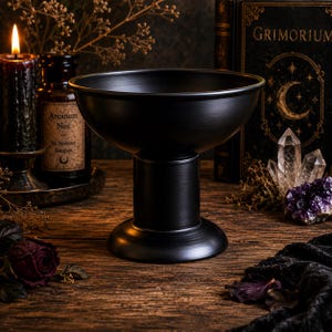 May include: A matte black pedestal bowl with a wide, shallow basin and a sturdy cylindrical base. The bowl features a smooth, uniform finish and a flared rim, standing upright as a singular decorative vessel for holding various small items.