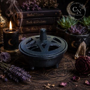 Könnte beinhalten: Ein Räuchergefäß aus schwarzem Gusseisen mit einem ausgeschnittenen Pentagramm-Design auf dem Deckel. Das runde Gefäß mit drei Standfüßen hat einen zentralen Griff auf dem abnehmbaren Deckel. Die Oberfläche ist texturiert und der Deckel lässt Rauch entweichen.