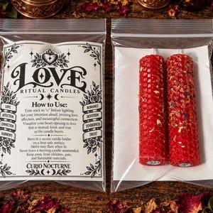 May include: Two red cylindrical beeswax candles with a honeycomb texture and embedded dried herbs, packaged in a clear plastic bag alongside a white instructional card titled Love Ritual Candles by Curio Nocturne with text instructions.