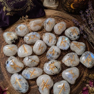 May include: A collection of polished, translucent white moonstone runes, each featuring a unique gold-engraved symbol. The stones are arranged in a cluster, displaying natural inclusions and iridescent blue flashes across their smooth, rounded surfaces.