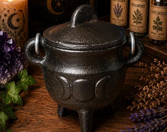May include: A black cast iron cauldron with a matching lid, featuring a triple moon symbol embossed on the front. The pot has three sturdy legs and two side handles with silver-toned rings attached, providing a textured and durable appearance.