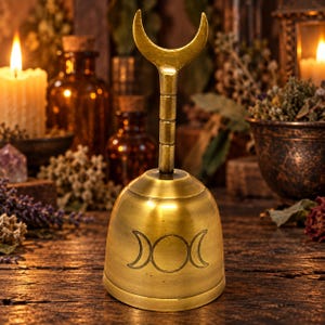May include: A brass bell featuring a crescent moon handle and a triple moon symbol engraved on the body. The bell has a smooth, metallic finish and a flared base, standing upright with a vertical stem connecting the handle to the main bell body.