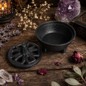 May include: A black cast iron cauldron with three small legs and a matching circular lid featuring a cut-out starburst pattern. The cauldron has a textured, matte finish and a deep, open interior, presented alongside its removable vented cover.