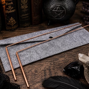 May include: Two L-shaped copper dowsing rods with smooth, metallic surfaces. The rods are positioned on top of a grey felt envelope-style carrying case that features a single black snap button closure at the center of the flap.