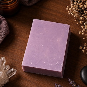 May include: A rectangular, lavender-colored bar of soap sits on a dark wooden surface. Surrounding the soap are a lit candle in a glass jar, a folded towel, crystals, dried flowers, a black stone, and sprigs of lavender.