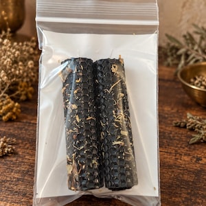 May include: Two cylindrical black beeswax candles with a textured honeycomb pattern and embedded dried botanical pieces, sealed inside a clear plastic zip-top bag with a white paper backing insert for stability and protection during storage.