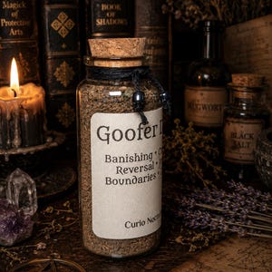 Puede incluir: Un frasco de vidrio transparente lleno de material granular marrón, sellado con un tapón de corcho. Una etiqueta blanca dice Goofer Dust, Banishing, Reversal, Boundaries y Curio Nocturna. Un cordel negro con cuentas oscuras rodea el cuello.