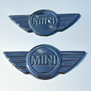 Peut inclure: Deux emblèmes MINI chromés. Chaque emblème présente le mot « MINI » dans un badge circulaire, avec des ailes stylisées s'étendant vers l'extérieur. Les emblèmes sont placés sur un fond clair et réfléchissant.