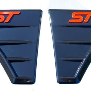 May include: Two dark blue car vent covers with the letters "ST" in orange. The covers have a triangular shape with horizontal lines. The background is white.