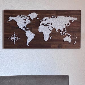 Könnte beinhalten: Eine Holz-Weltkarte mit dunkelbraunem Finish, auf der die Kontinente mit silbernem Faden umrissen sind. Eine silberne Windrose befindet sich in der linken unteren Ecke. Die Karte ist an einer Wand über einem grauen Sofa angebracht.