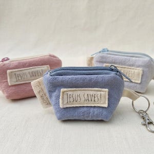 May include: Three small fabric zip pouches in dusty pink, periwinkle blue, and light grey. Each pouch features a rectangular cream-coloured fabric label stitched to the front with the text "JESUS SAVES!" and a metal lobster clasp keychain.