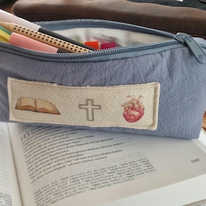 Könnte beinhalten: Ein schieferblaues Federmäppchen aus Stoff mit Reißverschluss, das mehrere Stifte enthält. Ein rechteckiger beiger Aufnäher auf der Vorderseite zeigt gedruckte Symbole eines offenen Buches, eines Kreuzes und eines roten Herzens. Ein kleines Etikett zeigt LORK'S.
