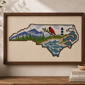 May include: A framed cross-stitch embroidery depicting the state of North Carolina. The design features blue mountains, green pine trees, a red cardinal bird on a branch, a black and white lighthouse, and blue ocean waves on a beige fabric background.