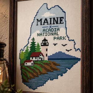 May include: Framed cross-stitch art depicting the state of Maine, featuring a lighthouse, trees, and the text "MAINE ACADIA NATIONAL PARK." The artwork is in a dark wooden frame, showcasing a coastal landscape with blue, green, and brown hues.