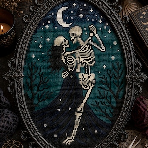 Puede incluir: Obra de arte de punto de cruz en un marco negro adornado. La imagen representa dos esqueletos bailando bajo una luna creciente y estrellas, sobre un fondo verde azulado y azul marino. El marco tiene forma ovalada.