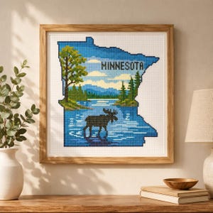 Puede incluir: Un bordado de punto de cruz cuadrado en un marco de madera clara que representa el estado de Minnesota. El diseño muestra un lago azul, la silueta negra de un alce, árboles verdes y un cielo azul claro. La palabra MINNESOTA está bordada en negro.