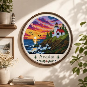 May include: A circular cross-stitch embroidery in a dark wood frame depicting a coastal lighthouse scene at sunset. The design features a white lighthouse with a red roof, rocky cliffs, pine trees, a vibrant purple and orange sky, and the text Acadia.