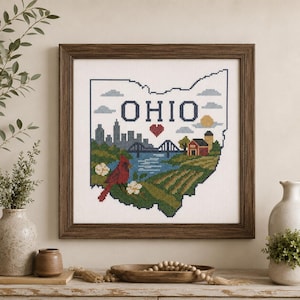 Pode incluir: Um bordado em ponto cruz emoldurado representando o estado de Ohio. O design apresenta um horizonte urbano, uma ponte sobre a água, um celeiro vermelho, um pássaro cardeal vermelho e flores brancas, com a palavra OHIO e um coração vermelho.