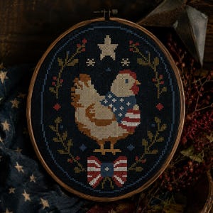 May include: Cross-stitch embroidery of a chicken wearing an American flag-themed scarf and bow tie, set against a dark blue background. The design includes a star, floral elements, and is framed in a wooden hoop. A patriotic home decor item.