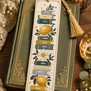May include: A rectangular cross-stitch bookmark featuring a vertical stack of books in shades of blue, green, and cream, interspersed with yellow lemons and white daisy flowers. The bookmark is finished with a cream-colored cord and tassel.