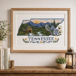 Könnte beinhalten: Ein gerahmtes Kreuzstichbild, das den Bundesstaat Tennessee darstellt, mit einer Landschaft aus Bergen, einer Wassermühle, einem Fluss, einem Vogel und Blumen. Das Wort TENNESSEE ist in blauen Buchstaben unter der Karte gestickt, flankiert von gelben Sternen.