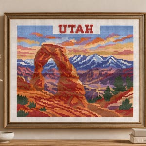 May include: A framed cross-stitch embroidery depicting a landscape of the Utah desert with a prominent red rock arch, distant blue mountains, and a sunset sky. The word UTAH is stitched in red capital letters at the top center of the artwork.