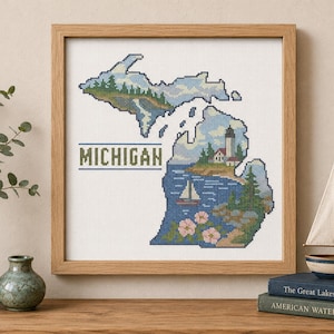 May include: A square framed cross-stitch embroidery depicting the state of Michigan. The design features a lighthouse, a sailboat, pine trees, and pink flowers within the state outline. The word MICHIGAN is stitched in dark green block letters.