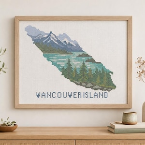 May include: A cross-stitch embroidery art piece in a light wood frame depicting a landscape of Vancouver Island. The design features snow-capped mountains, a turquoise lake, and dark green pine trees, with the text VANCOUVER ISLAND below.