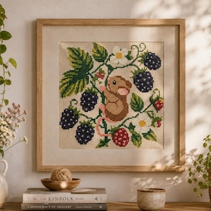 Puede incluir: Un bordado en punto de cruz enmarcado de forma cuadrada que muestra un ratón marrón trepando entre hojas verdes, flores blancas y moras de color púrpura oscuro sobre un fondo de tela beige claro. La obra está en un marco de madera clara.