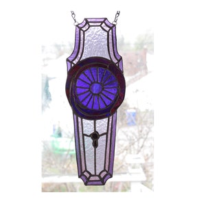 May include: A stained glass window panel in shades of purple and lavender. The panel features a circular design with radiating lines, a keyhole detail, and a textured background. It is suspended by a chain.