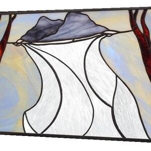 May include: Stained glass panel featuring a landscape scene. The artwork depicts two dark gray mountains, white flowing shapes, and two red-brown tree trunks. The background has a mix of light blue, yellow, and white colors, all framed in a black border.