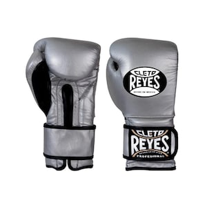 Peut inclure: Paire de gants de boxe argentés avec des détails noirs. Les gants arborent le logo "CLETO REYES" en noir et blanc sur le poignet et le dos. Fabriqués en cuir, ces gants sont conçus pour la boxe professionnelle.