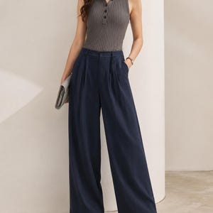 Puede incluir: Un top sin mangas de punto acanalado gris con cuello polo y cierre de botones, combinado con pantalones anchos azul marino. El conjunto se completa con zapatos negros de punta y un clutch gris.
