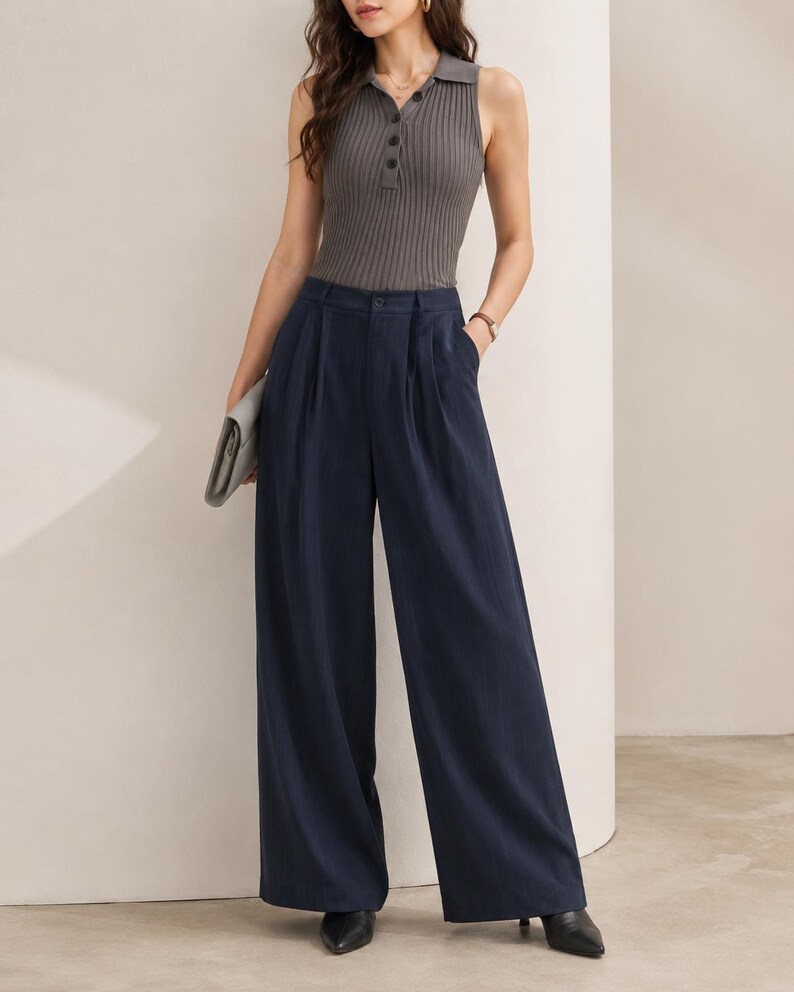 Puede incluir: Un top sin mangas de punto acanalado gris con cuello polo y cierre de botones, combinado con pantalones anchos azul marino. El conjunto se completa con zapatos negros de punta y un clutch gris.