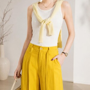 Puede incluir: Un conjunto femenino que incluye una camiseta sin mangas blanca, un su&eacute;ter amarillo atado alrededor del cuello y pantalones amarillos de cintura alta. El conjunto se complementa con un collar dorado y un reloj negro. Se sostiene un bolso crema en la mano izquierda.