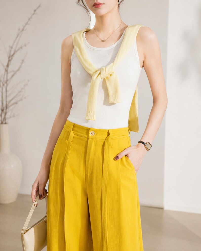 Puede incluir: Un conjunto femenino que incluye una camiseta sin mangas blanca, un su&eacute;ter amarillo atado alrededor del cuello y pantalones amarillos de cintura alta. El conjunto se complementa con un collar dorado y un reloj negro. Se sostiene un bolso crema en la mano izquierda.