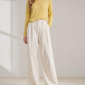 Puede incluir: Una persona con una camisa de manga larga de color amarillo mostaza y pantalones de pierna ancha color crema. Los pantalones tienen cintura alta y cintur&oacute;n a juego. El conjunto se completa con bailarinas amarillas. El fondo es una pared blanca lisa.