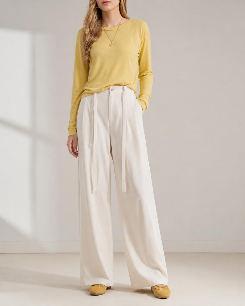 Puede incluir: Una persona con una camisa de manga larga de color amarillo mostaza y pantalones de pierna ancha color crema. Los pantalones tienen cintura alta y cintur&oacute;n a juego. El conjunto se completa con bailarinas amarillas. El fondo es una pared blanca lisa.