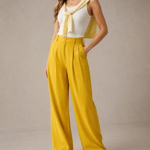 Puede incluir: Una mujer con una camiseta sin mangas blanca, un su&eacute;ter amarillo atado a los hombros y pantalones amarillos de cintura alta y pernera ancha. Los pantalones tienen cierre de bot&oacute;n y bolsillos. Zapatillas blancas completan el conjunto.