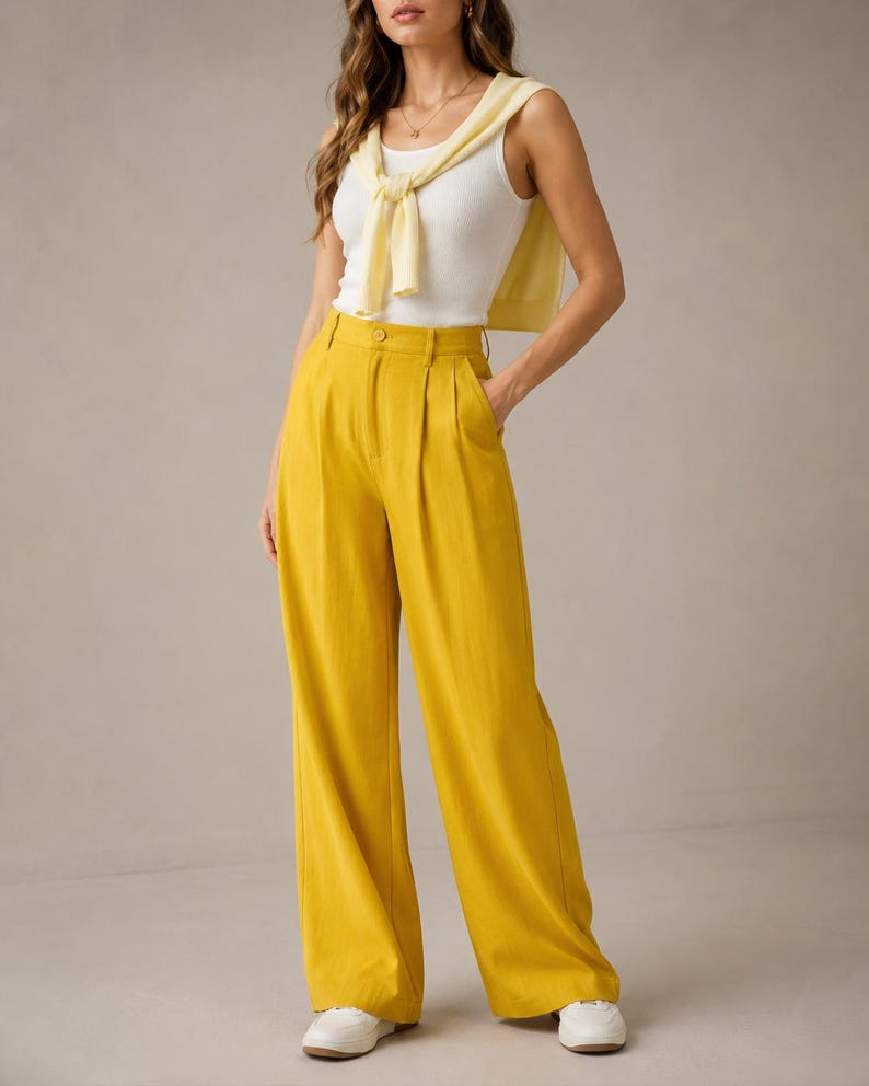 Puede incluir: Una mujer con una camiseta sin mangas blanca, un su&eacute;ter amarillo atado a los hombros y pantalones amarillos de cintura alta y pernera ancha. Los pantalones tienen cierre de bot&oacute;n y bolsillos. Zapatillas blancas completan el conjunto.