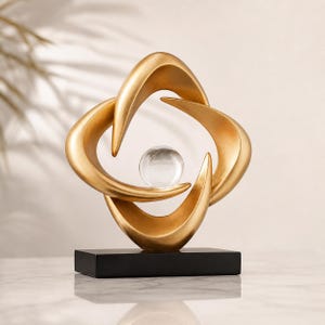 May include: A gold-colored abstract sculpture featuring four curved, interlocking shapes surrounding a clear glass sphere. The sculpture sits on a black rectangular base, creating a modern decorative piece.
