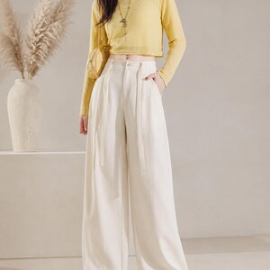 Puede incluir: Un conjunto femenino con un top corto amarillo de manga larga y pantalones de pierna ancha color crema. El conjunto se complementa con un peque&ntilde;o bolso amarillo y zapatos planos amarillos. Un jarr&oacute;n con plantas altas y secas en el fondo.